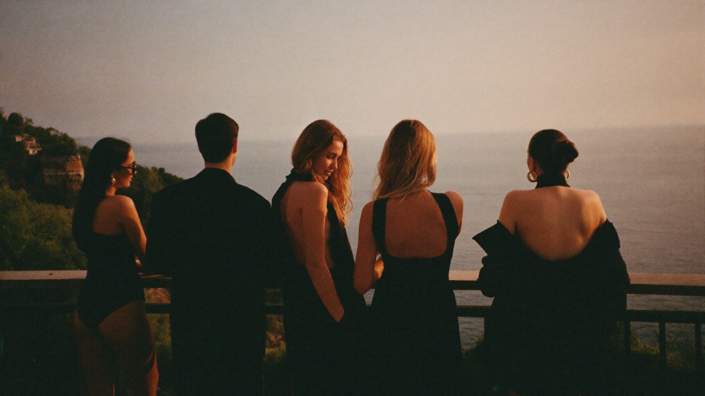 Friends in head-to-toe black at a Mediterranean clifftop terrace looking out to sea at sunset