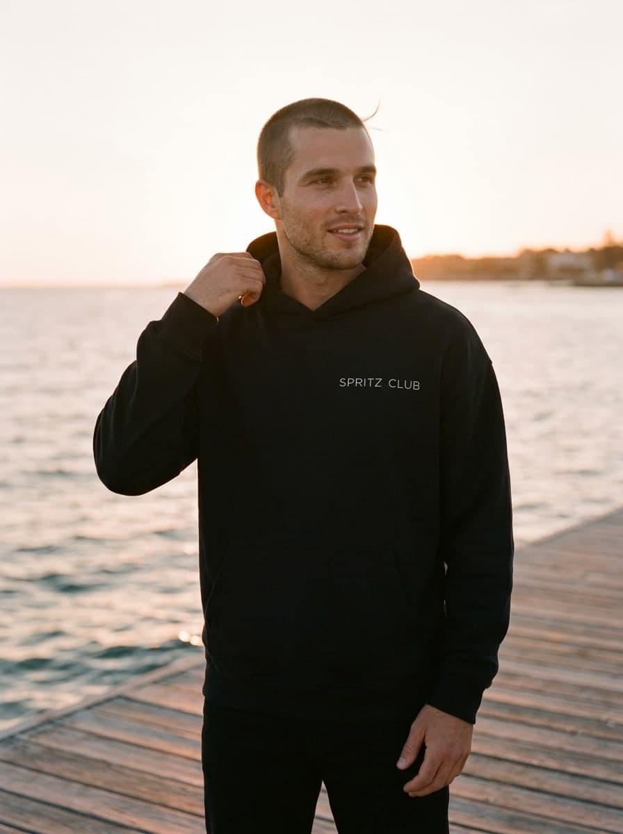 Man in the black Spritz Club heavyweight hoodie on a sunlit jetty by the sea at golden hour